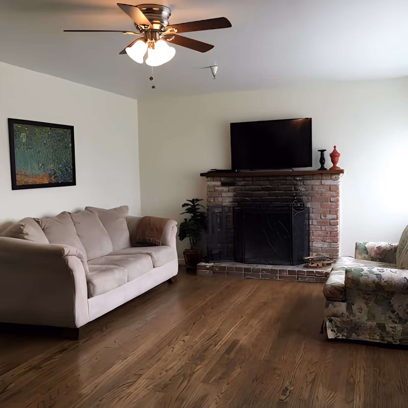 Living room with a sofa and armchair, hardwood floors, ceiling fan, and a brick fireplace topped by a TV.