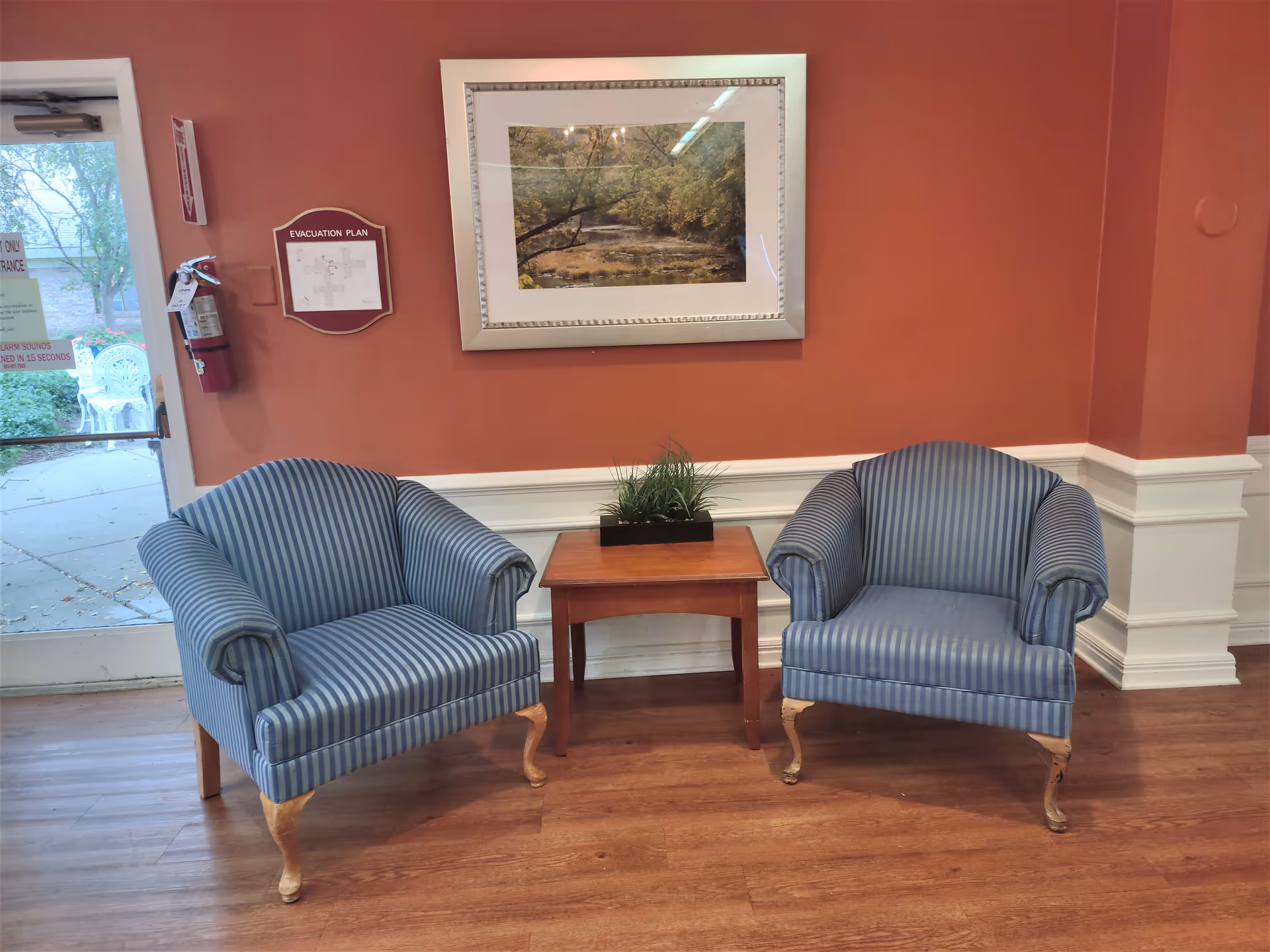 Two blue striped armchairs flanking a small wooden table with a plant beneath a framed landscape painting in a facility seating area.