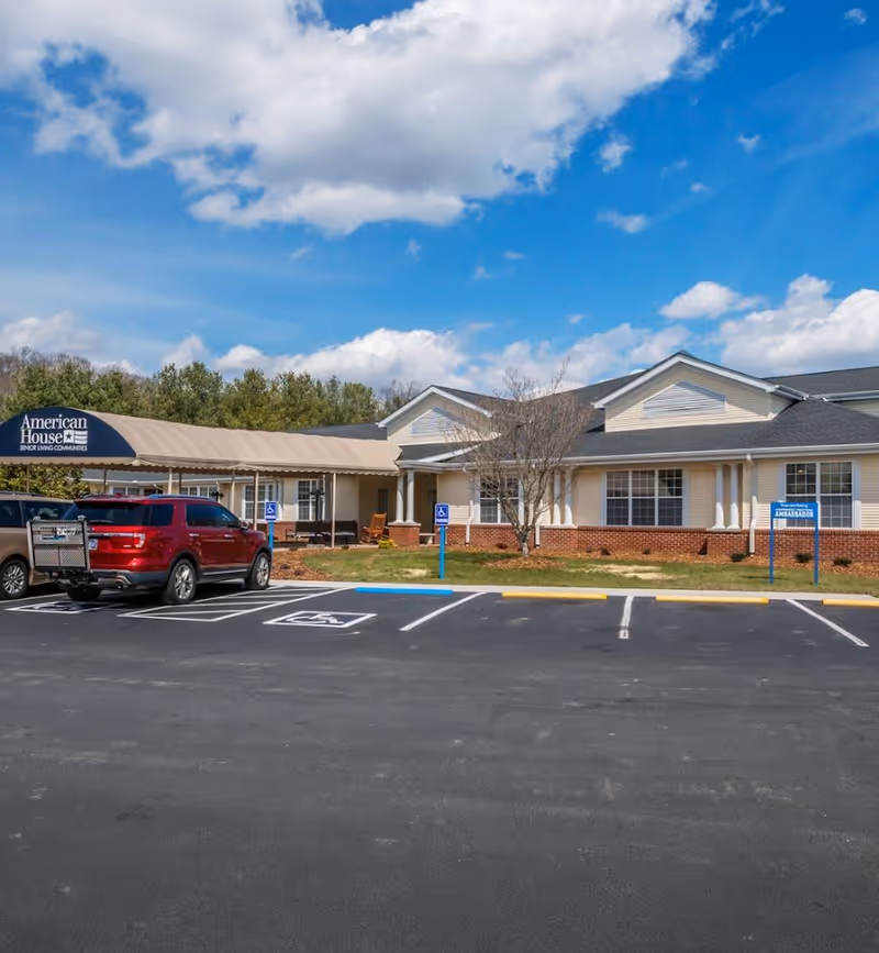 Exterior view of American House Johnson City senior living community building with a covered entrance, parked cars in front, and a clear blue sky with some clouds.