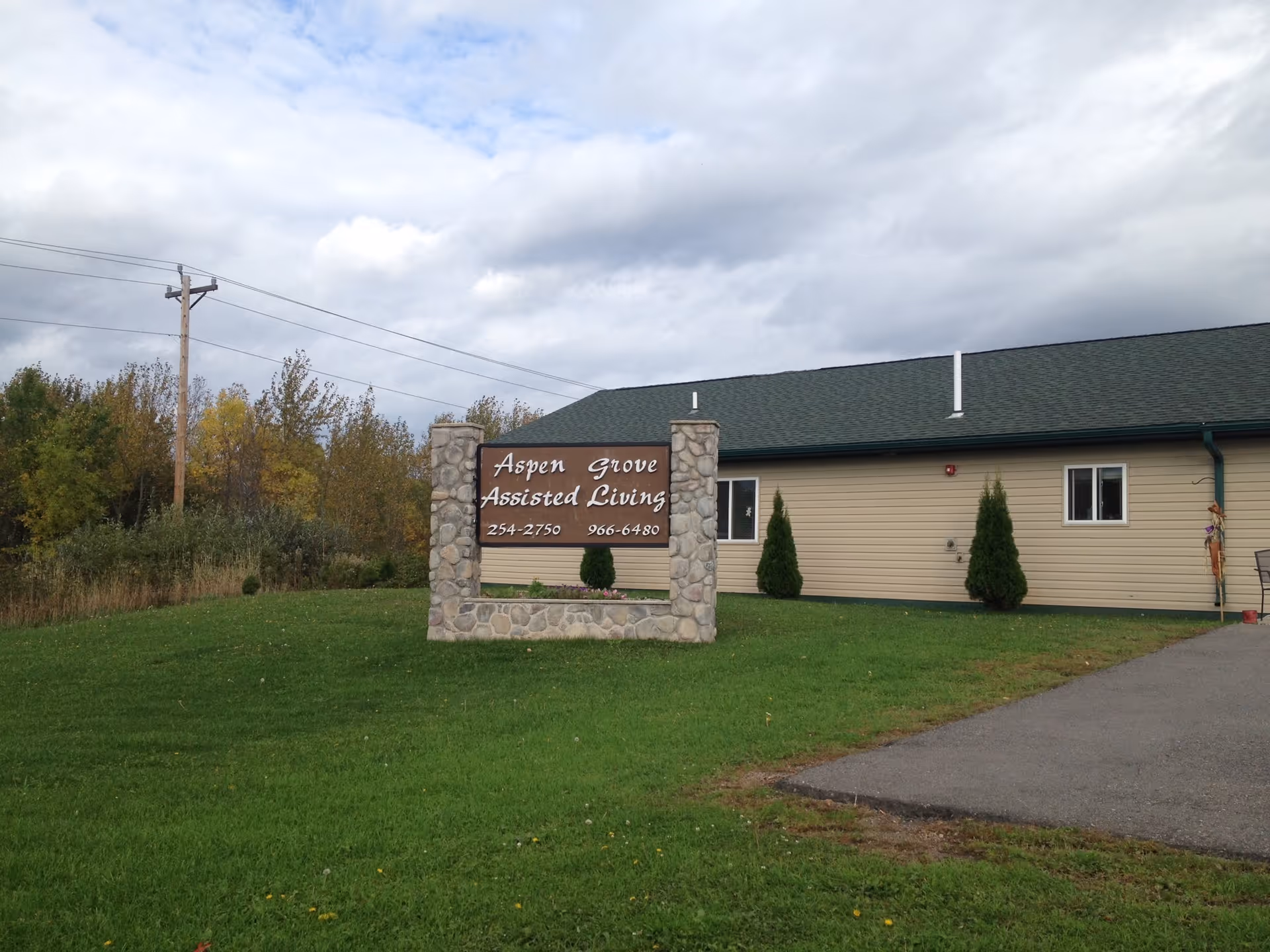 A stone-framed sign on a grassy lawn in front of a beige building with a green roof. The sign reads 'Aspen Grove Assisted Living' with two phone numbers below. Trees and a cloudy sky are visible in the background.
