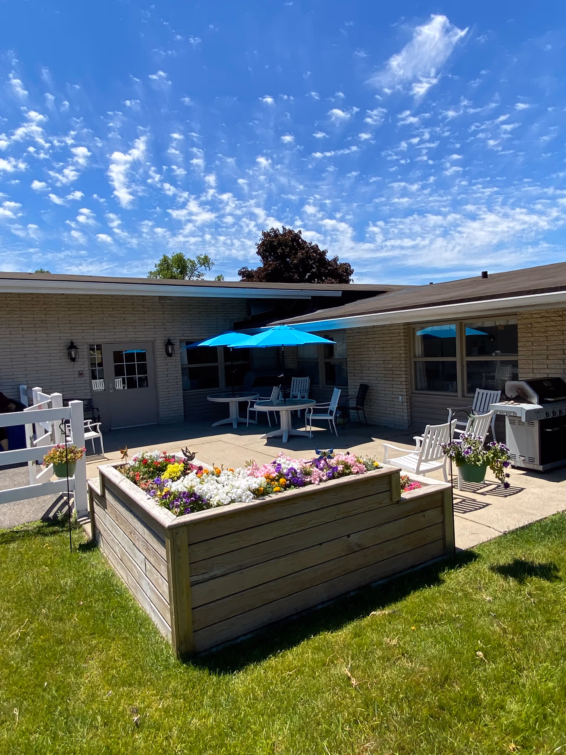 Outdoor patio area at Brickyard Healthcare - Valparaiso Care Center featuring a raised wooden flower bed filled with colorful flowers, several white chairs, tables with blue umbrellas, a barbecue grill, and a building with large windows under a bright blue sky with scattered clouds.