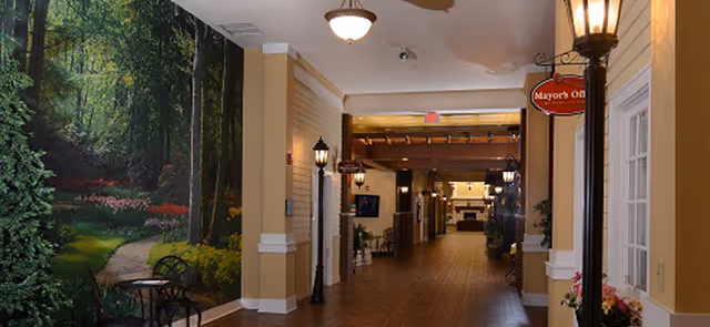 A wide indoor hallway with warm lighting and a wooden floor. On the left side, there is a large mural depicting a forest scene with a path, trees, and flowers. There are black streetlamp-style lights along the hallway and a small table with chairs near the mural. On the right side, there is a window and a sign hanging from a streetlamp that reads 'Mayor's Office.' The hallway extends into the distance with more lighting and furniture visible.