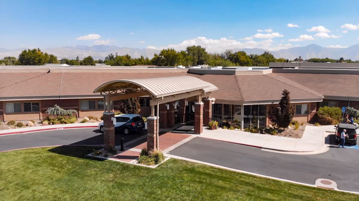 Exterior view of Rocky Mountain Care - Hunter Hollow facility showing the main entrance with a covered drop-off area, a car parked under the canopy, landscaped greenery, and mountains in the background under a blue sky with scattered clouds.