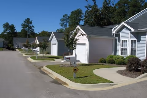 A row of single-story residential buildings with attached garages along a paved street. Each unit has a small front yard with green grass, bushes, and a mailbox. Trees and clear blue sky are visible in the background.