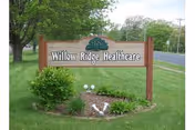 Wooden Willow Ridge Healthcare sign on a grassy lawn with trees and road in background.