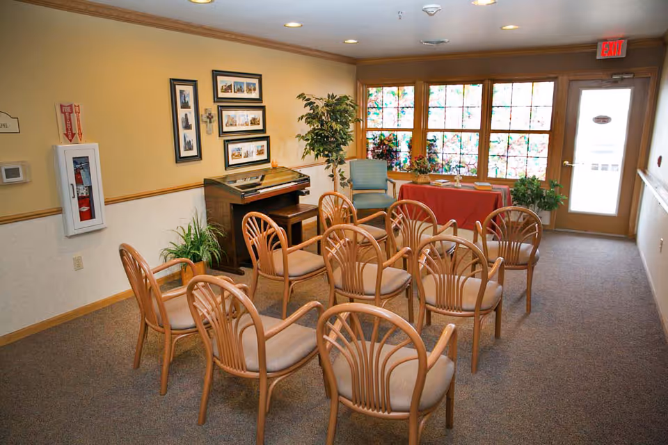 A senior living facility common room with wooden chairs, plants, a piano, framed photos, and windows.