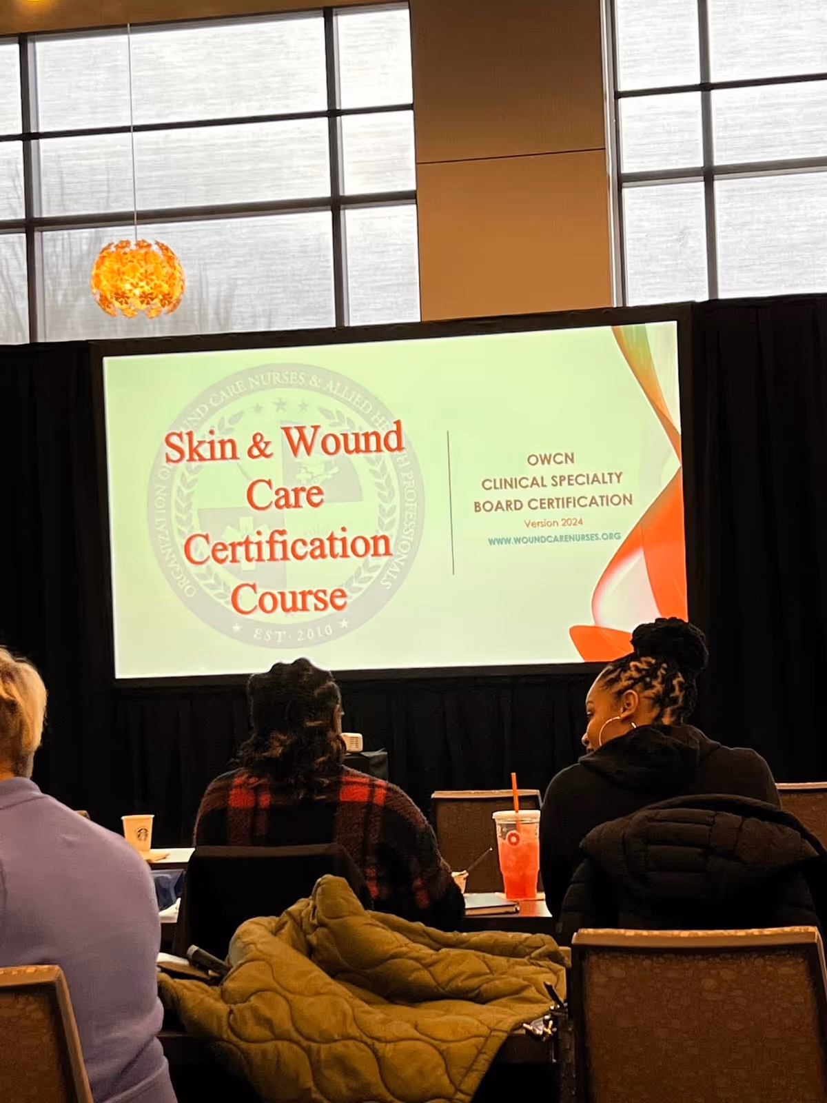 Conference room with a large screen displaying Skin and Wound Care Certification Course; attendees seated.