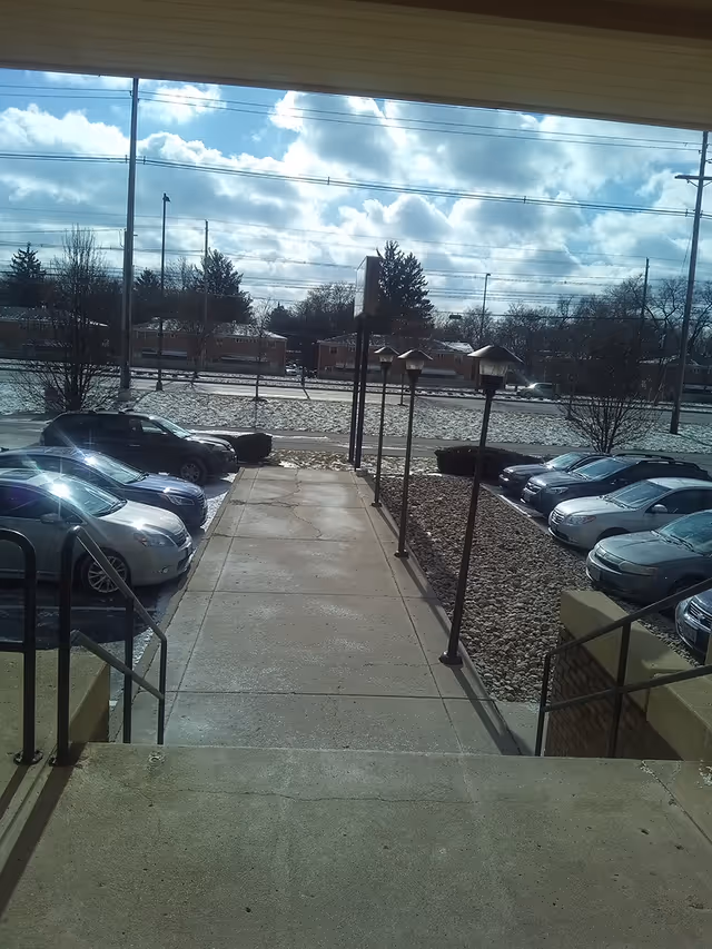 Exterior view of a facility entrance with parked cars, stairs, lamps, and a flooded street.