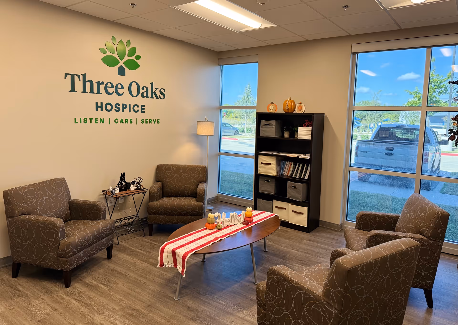 Three Oaks Hospice lobby with chairs, a coffee table, bookshelf, and large windows.