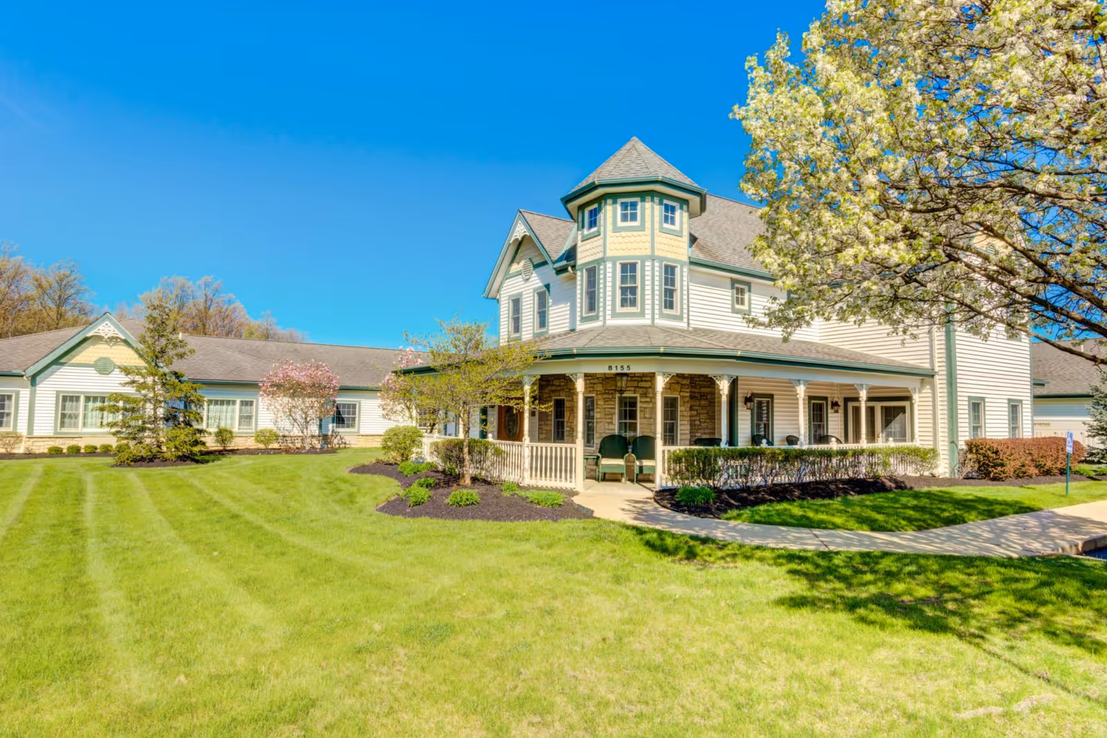 Exterior view of a large, two-story senior living facility building with white siding and green trim, featuring a wraparound porch with chairs. The building is surrounded by a well-maintained lawn, landscaped bushes, and blooming trees under a clear blue sky.
