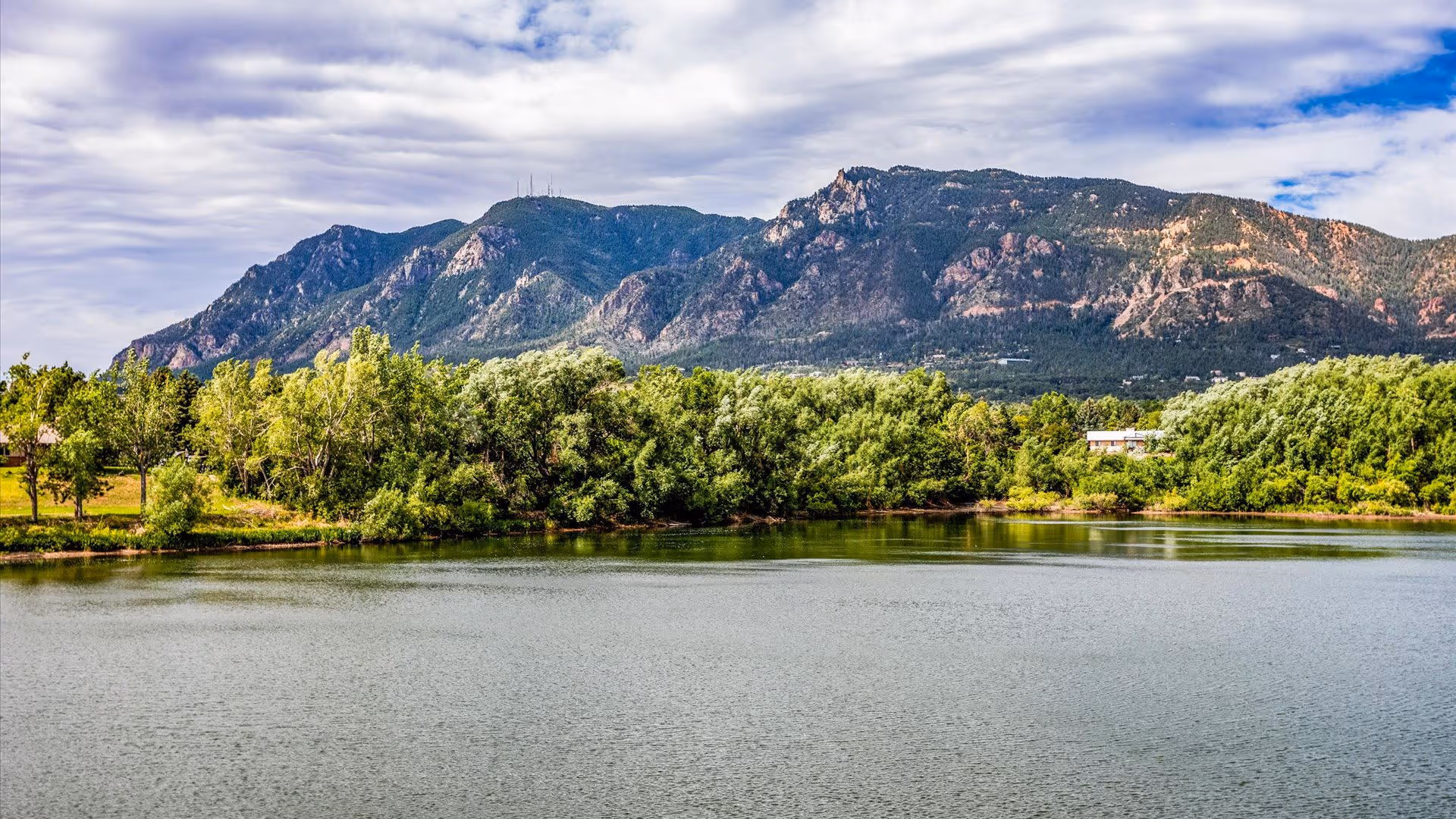 A scenic view of a calm lake with lush green trees along the shoreline and a large mountain range in the background under a partly cloudy sky.
