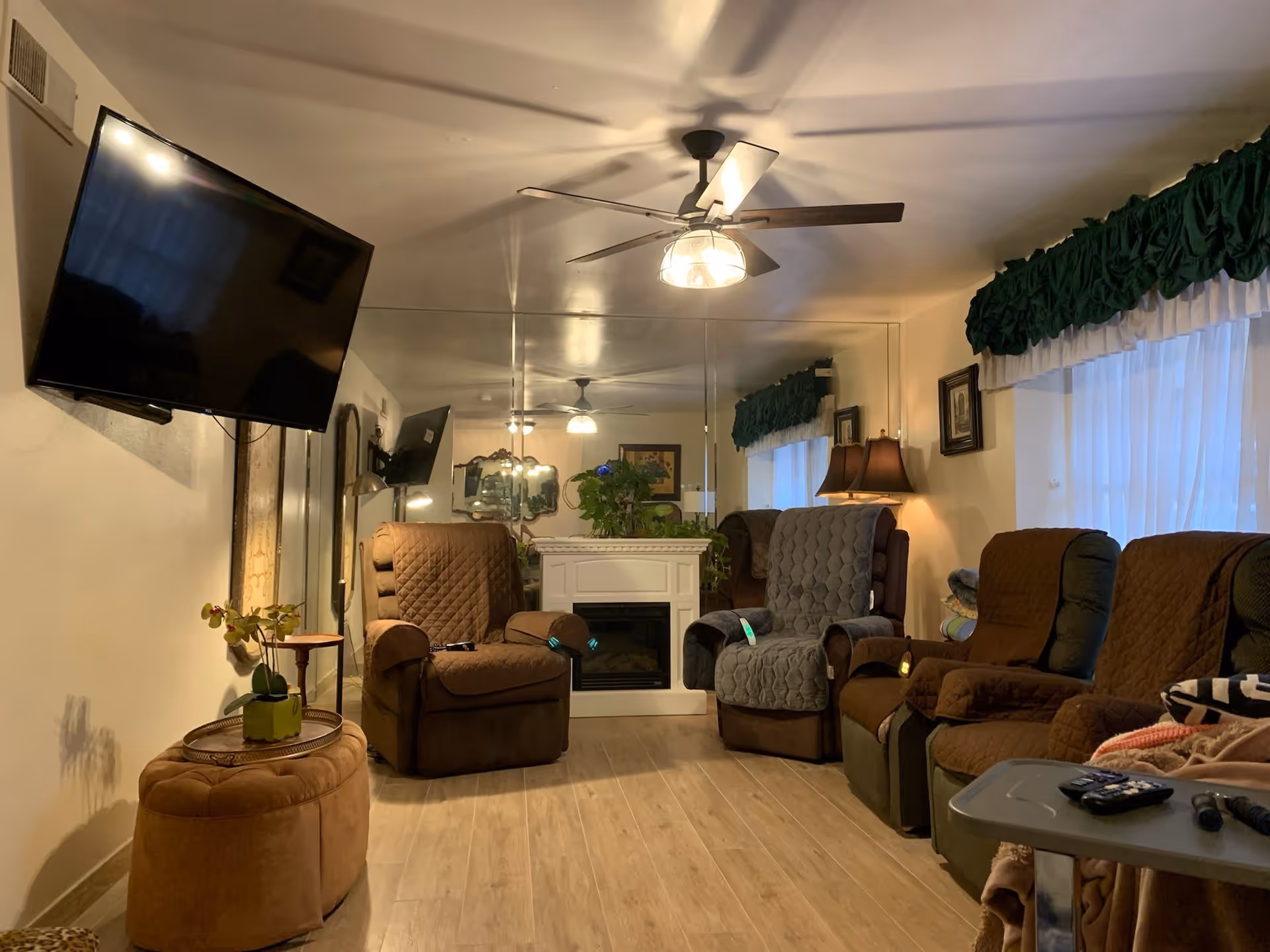 Cozy living room with multiple recliner chairs arranged around a faux fireplace, a wall-mounted TV, ceiling fan, and mirrored wall.