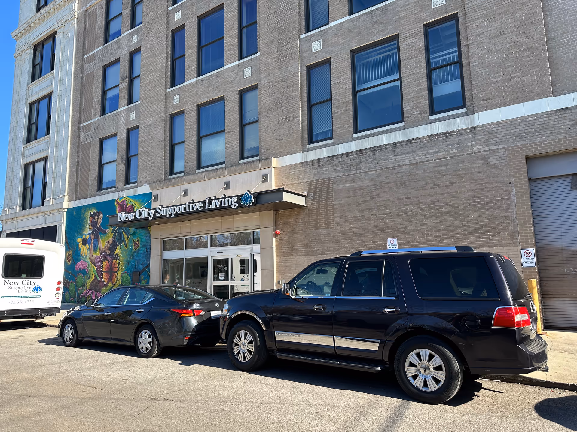 Street-level view of the New City Supportive Living building entrance with parked cars and a colorful mural.