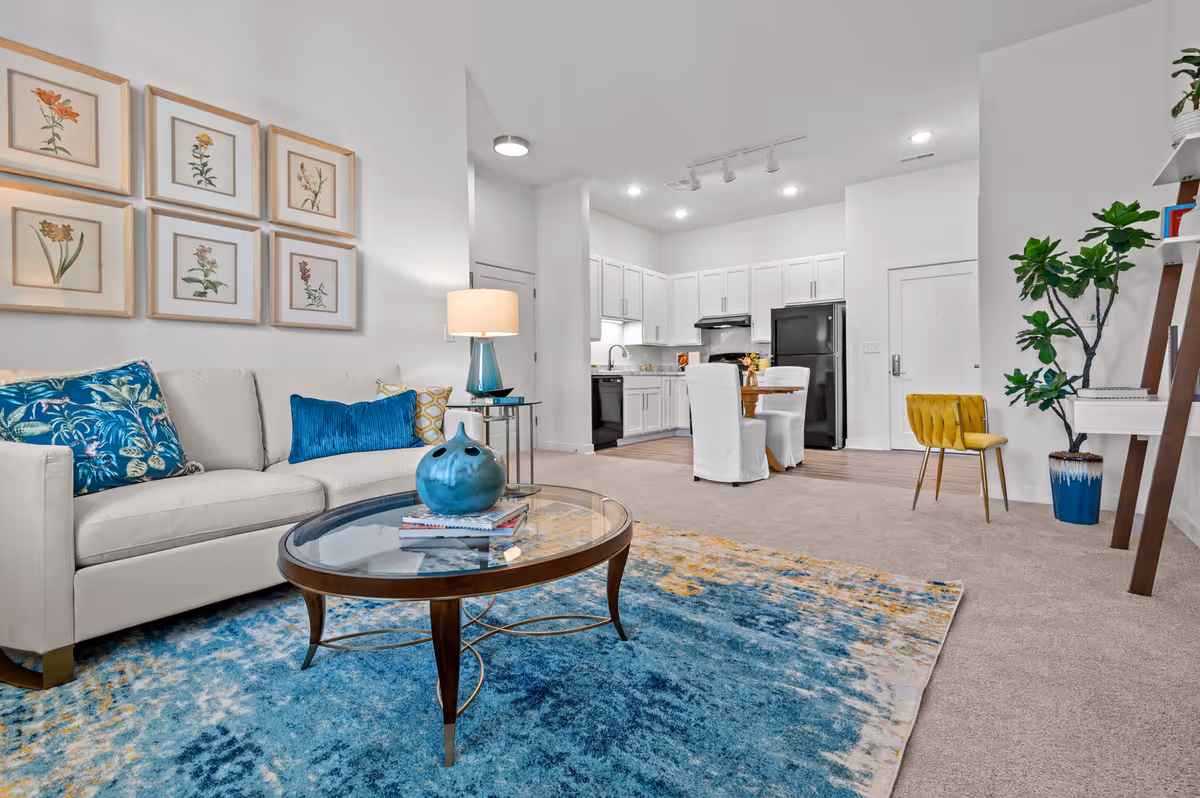 Bright open living room with sofa, coffee table, artwork, and a white kitchen in background.