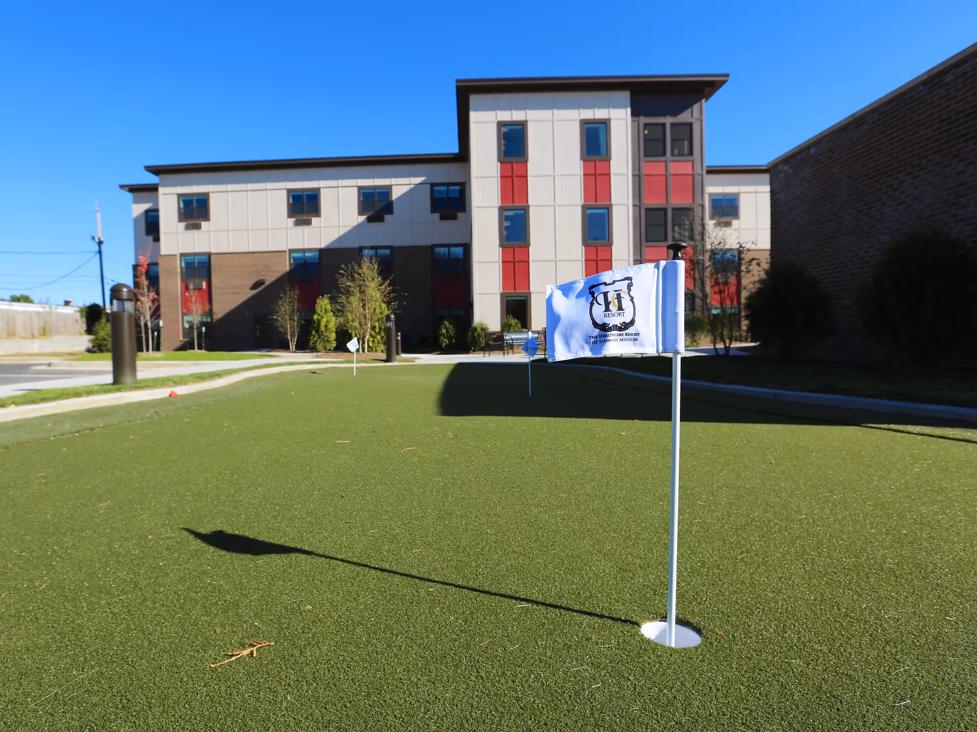 A putting green with small flags in front of a multi-story rehabilitation center building under a clear blue sky.