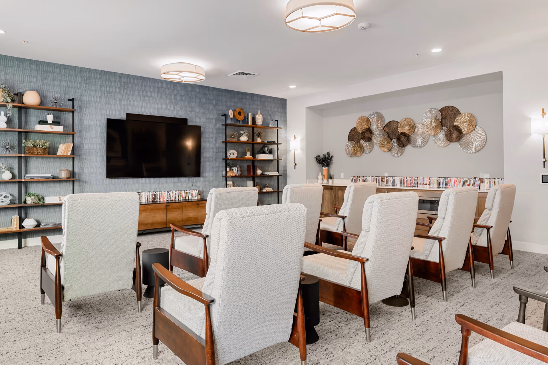 A cozy media room with multiple beige upholstered armchairs arranged in rows facing a large flat-screen TV mounted on a textured blue-gray wall. The room features wooden shelving units with decorative items and books, a wooden cabinet below the TV, and a wall adorned with circular metallic art pieces. The space is well-lit with ceiling lights and wall sconces.