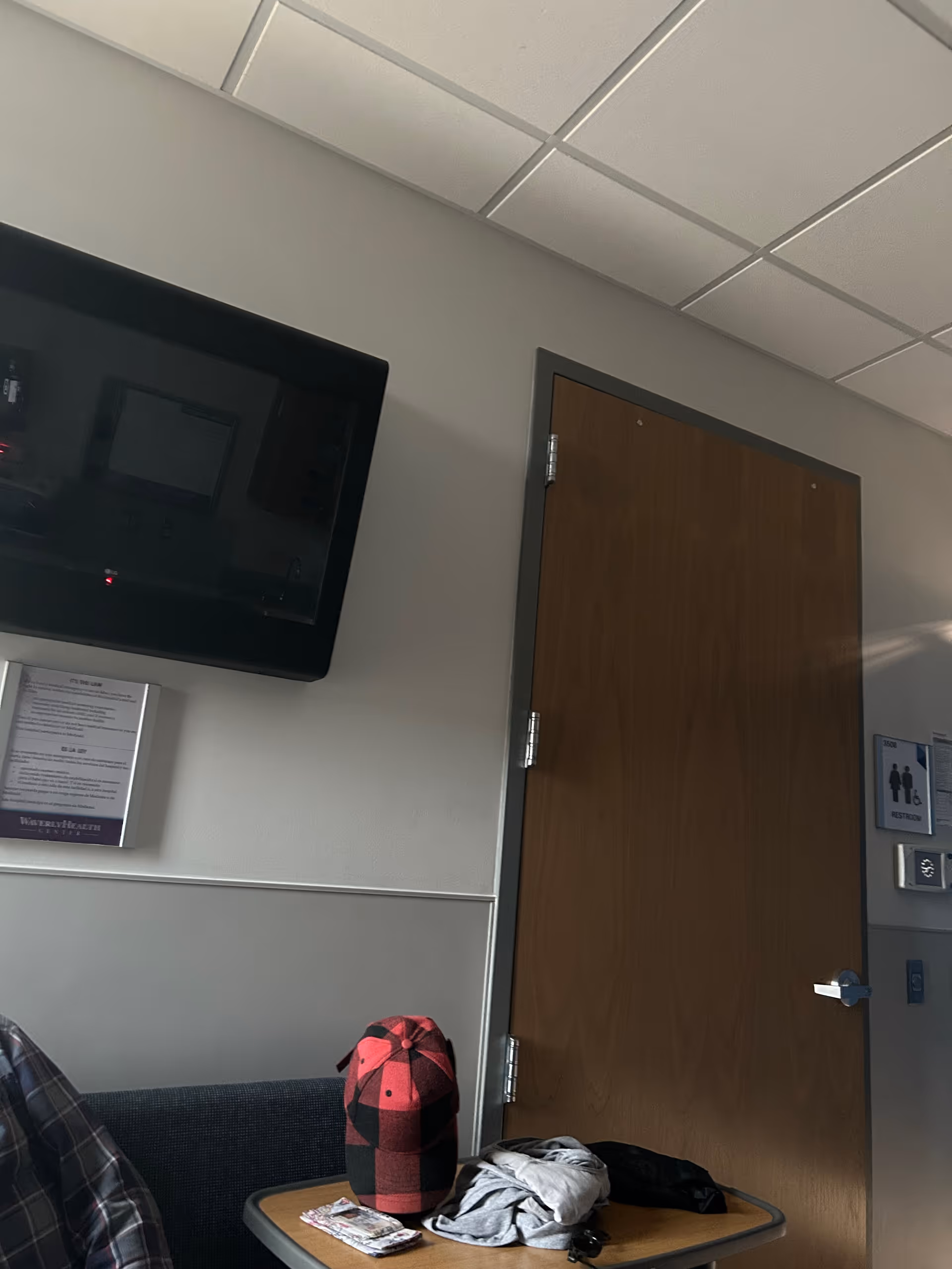 Interior waiting area with wall-mounted TV, wooden door, couch, and cluttered table.