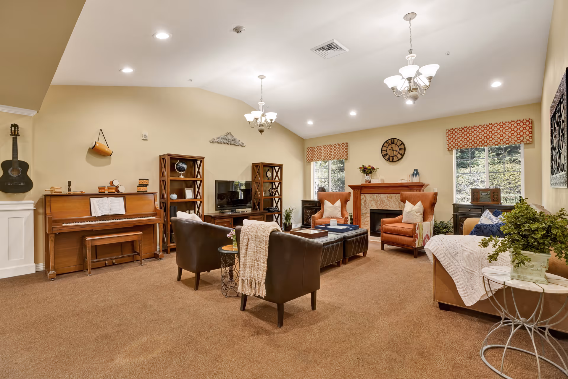 A cozy living room with beige walls and carpet, featuring a piano with a bench, a black guitar hanging on the wall, two black leather chairs with a small round table between them, two orange armchairs near a fireplace, a sofa with a white throw blanket, two windows with patterned valances, a wall clock above the fireplace, and decorative shelves with various items including a globe and framed pictures.