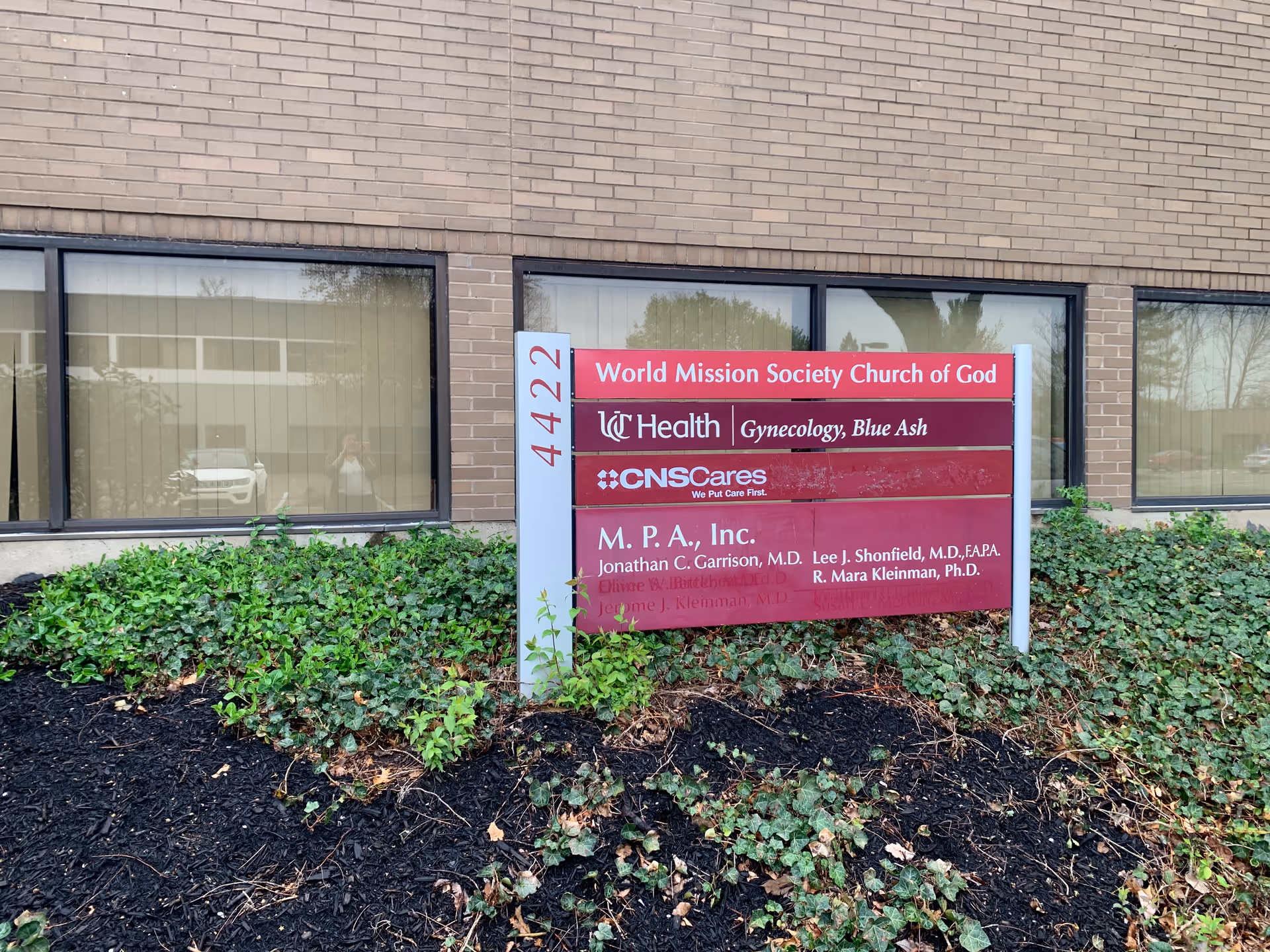 Front of a brick building with a multi-organization sign and surrounding shrubs.