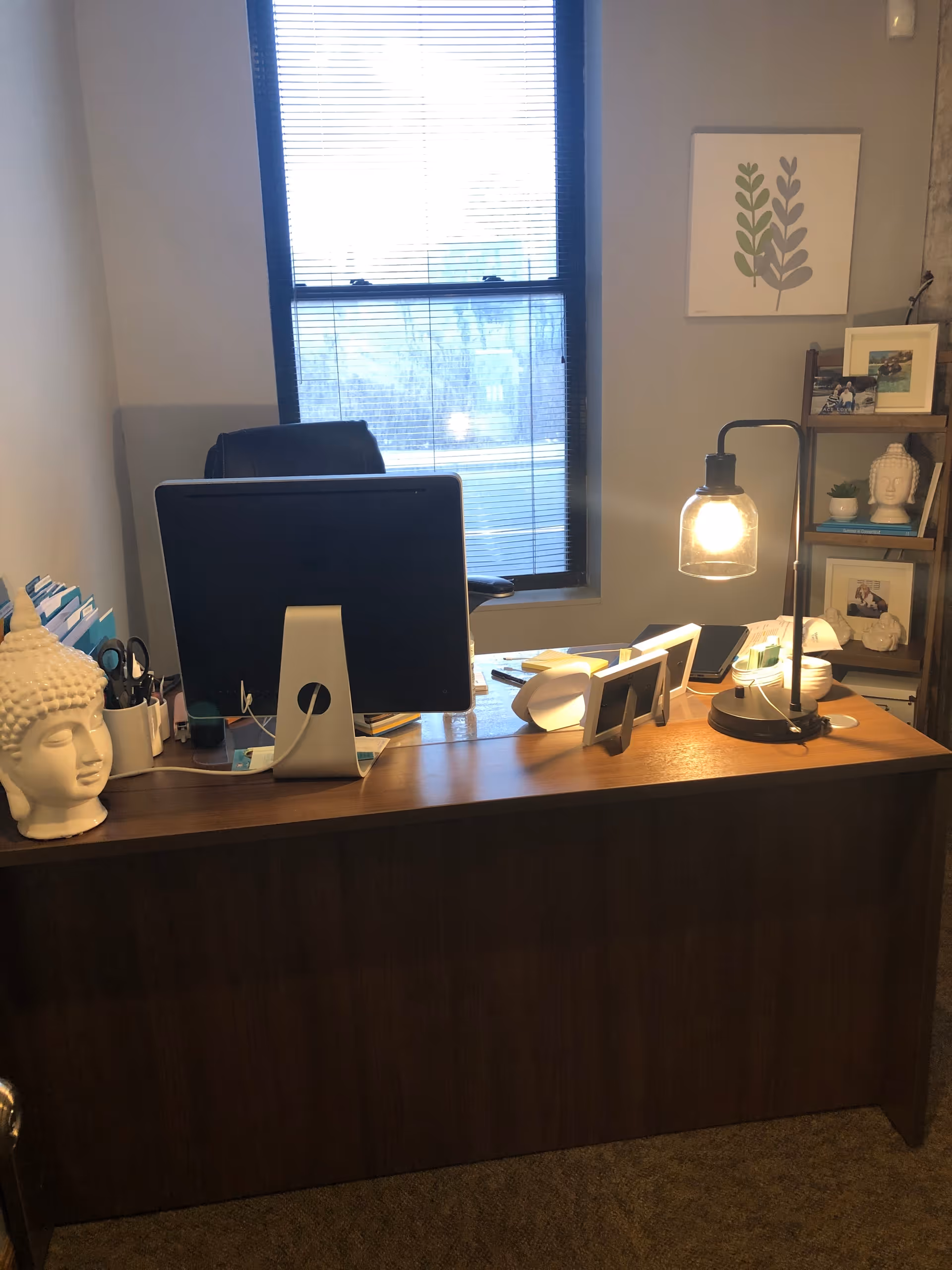 A neat office desk with computer, lamp, Buddha statue, and photo shelves by a window.