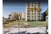 A multi-story beige senior living building with a manicured lawn and palm trees along a sidewalk.