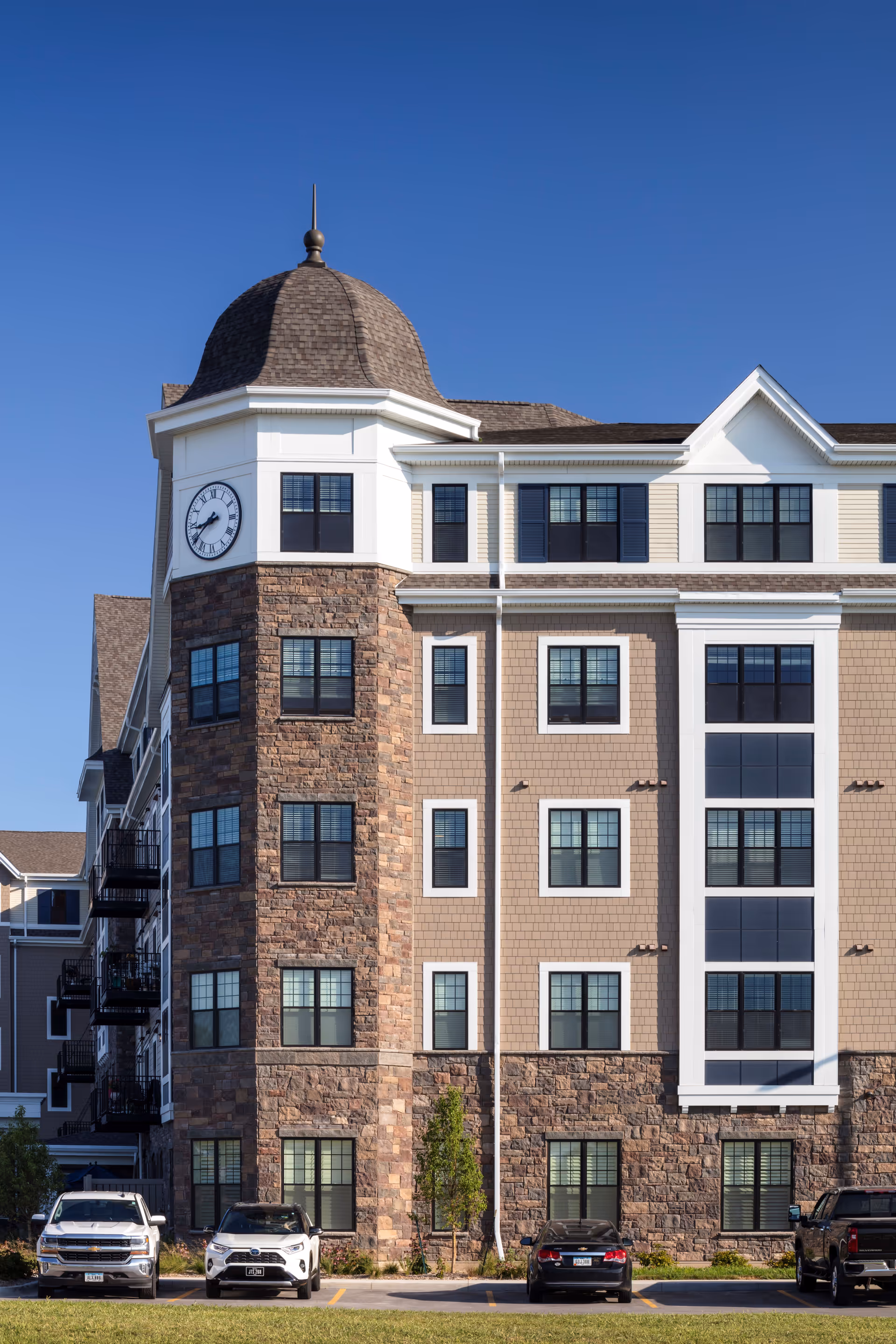 Front exterior of a multi-story senior living building with a clock tower, stone facade, and cars parked in front.