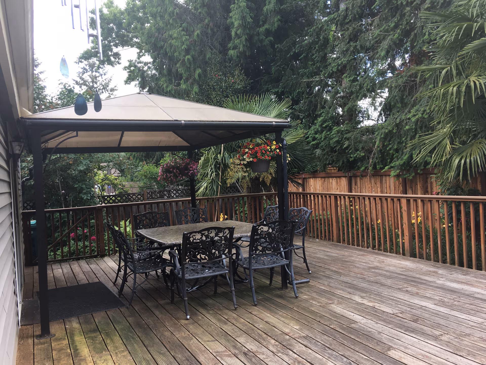 A wooden deck with a canopy-covered dining table and ornate chairs, surrounded by greenery.