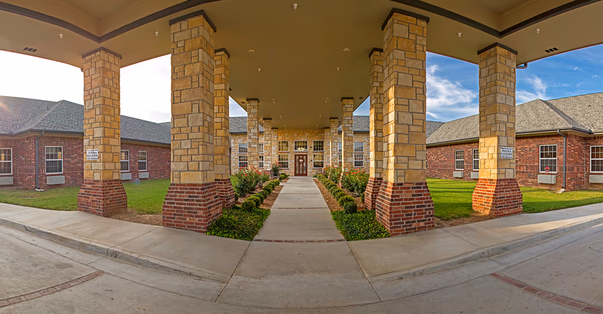 Covered entrance walkway to a brick and stone building with multiple stone pillars and landscaped flower beds on either side of the concrete path leading to a wooden door with windows.