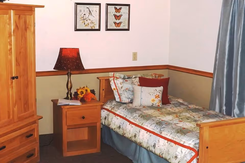 A cozy bedroom with a single wooden bed adorned with floral bedding and multiple pillows. Next to the bed is a wooden nightstand with a decorative lamp and a small bouquet of flowers. On the wall above the bed are two framed pictures featuring birds and butterflies. There is a wooden wardrobe on the left side and blue curtains on the right side of the room.