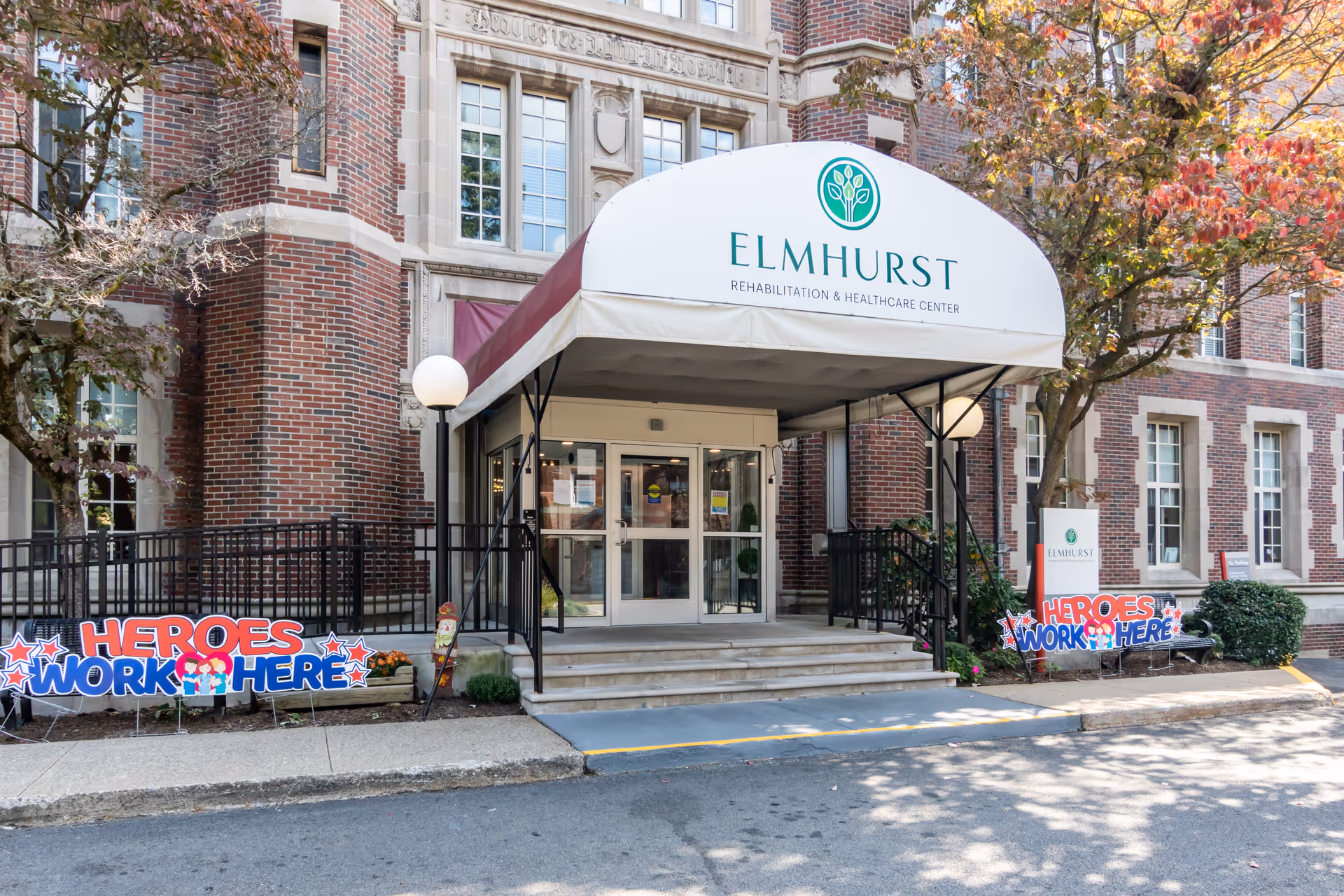 Elmhurst Rehabilitation & Healthcare Center entrance with white awning, brick building, and 'Heroes Work Here' signs.