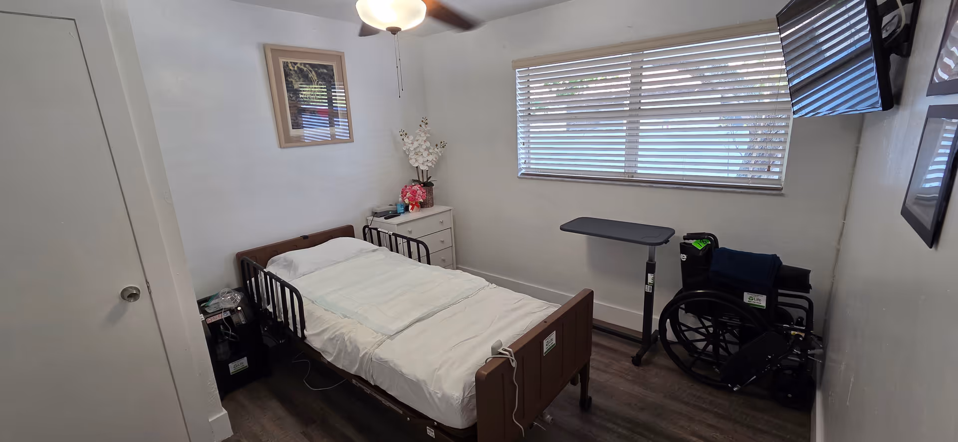 Small bedroom in a senior facility with a single bed, dresser, wheelchair, and window blinds.