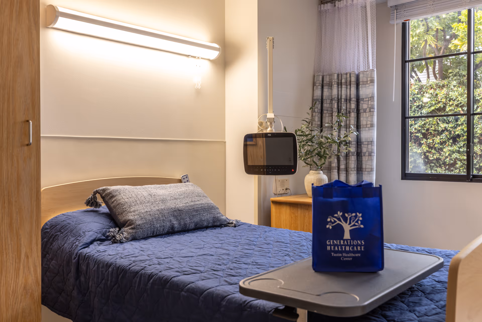 A hospital-style bedroom with blue quilted bed, window, plant, and a blue bag on a tray.