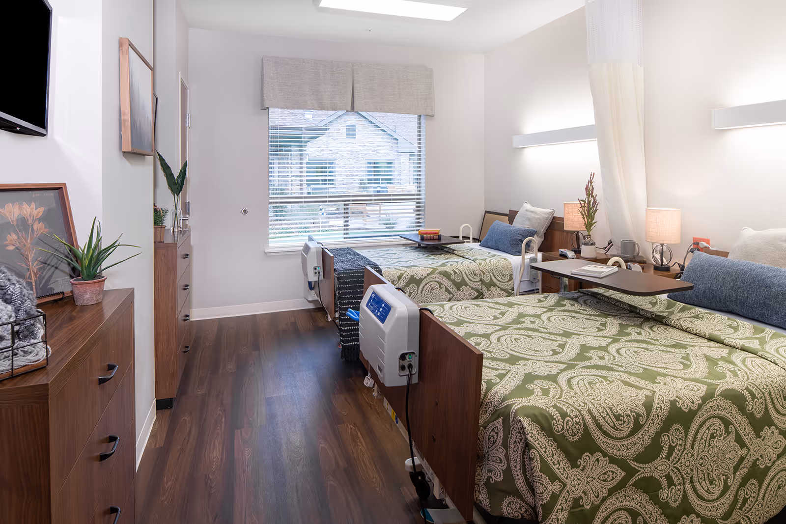 Two twin beds in a well-lit senior living bedroom with wood floors and plants.