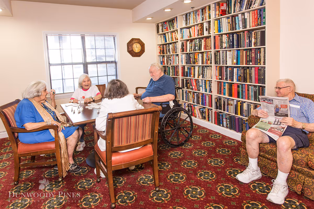 A group of elderly people in a cozy room with a large bookshelf filled with books. Four seniors are seated around a table playing a game, while another man sits on a patterned armchair reading a newspaper. The room has a red carpet with floral patterns and a window letting in natural light.