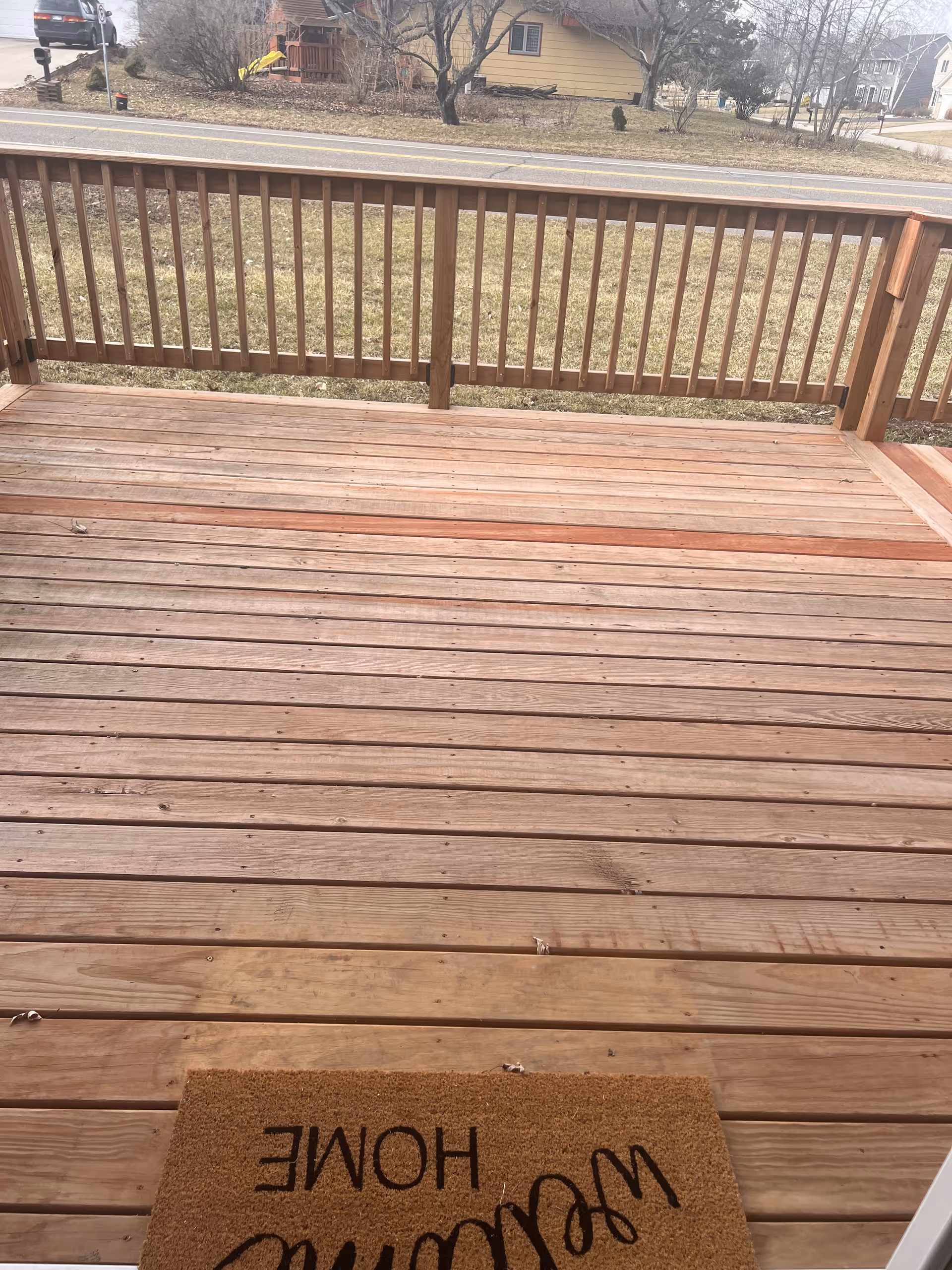 Wooden deck with railing and a Welcome Home mat overlooking a residential street.