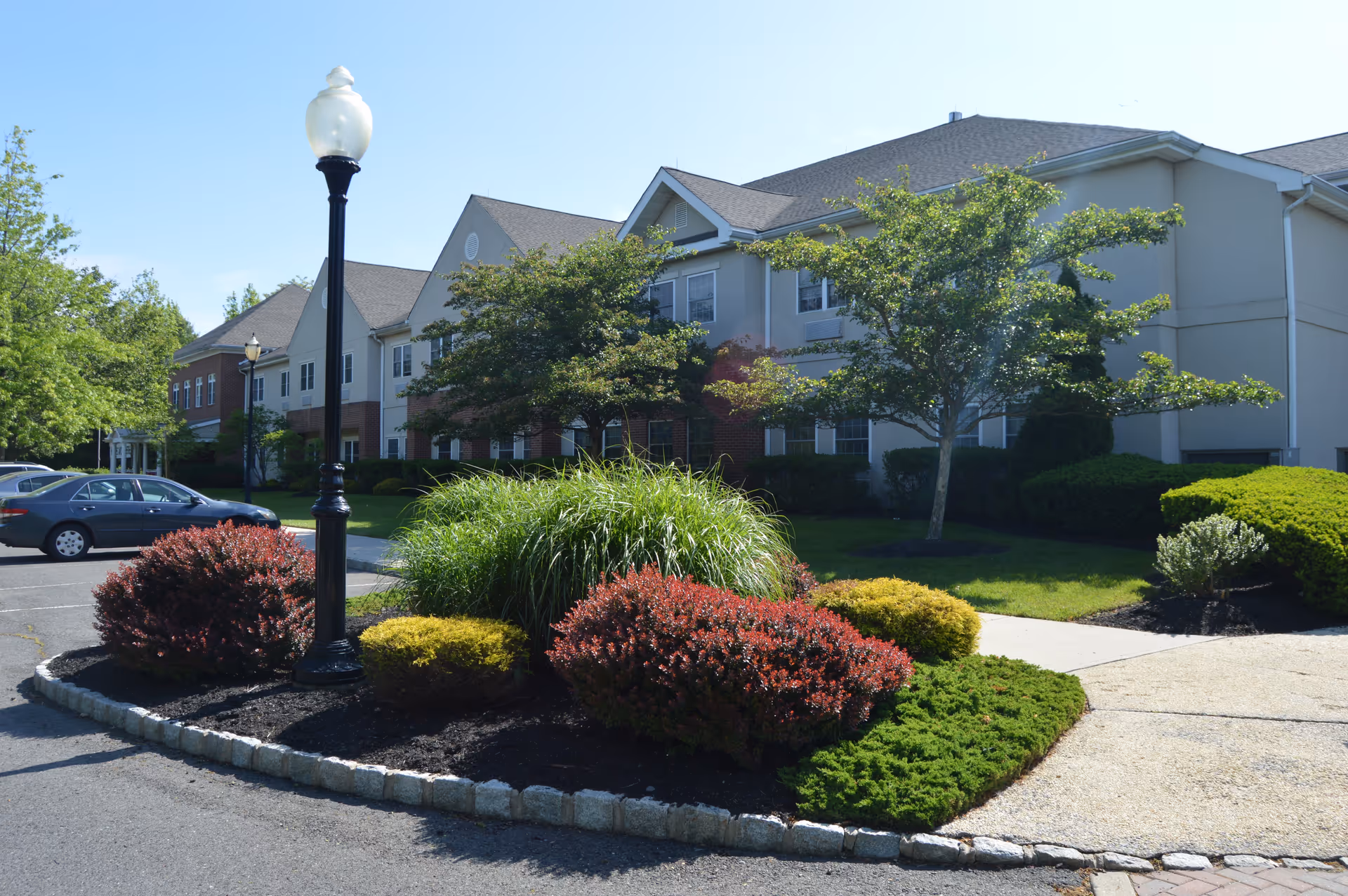 Exterior view of a senior living facility named Rose Hill with a landscaped garden area featuring various shrubs and a lamp post in the foreground, and a two-story building with multiple windows in the background under a clear blue sky.