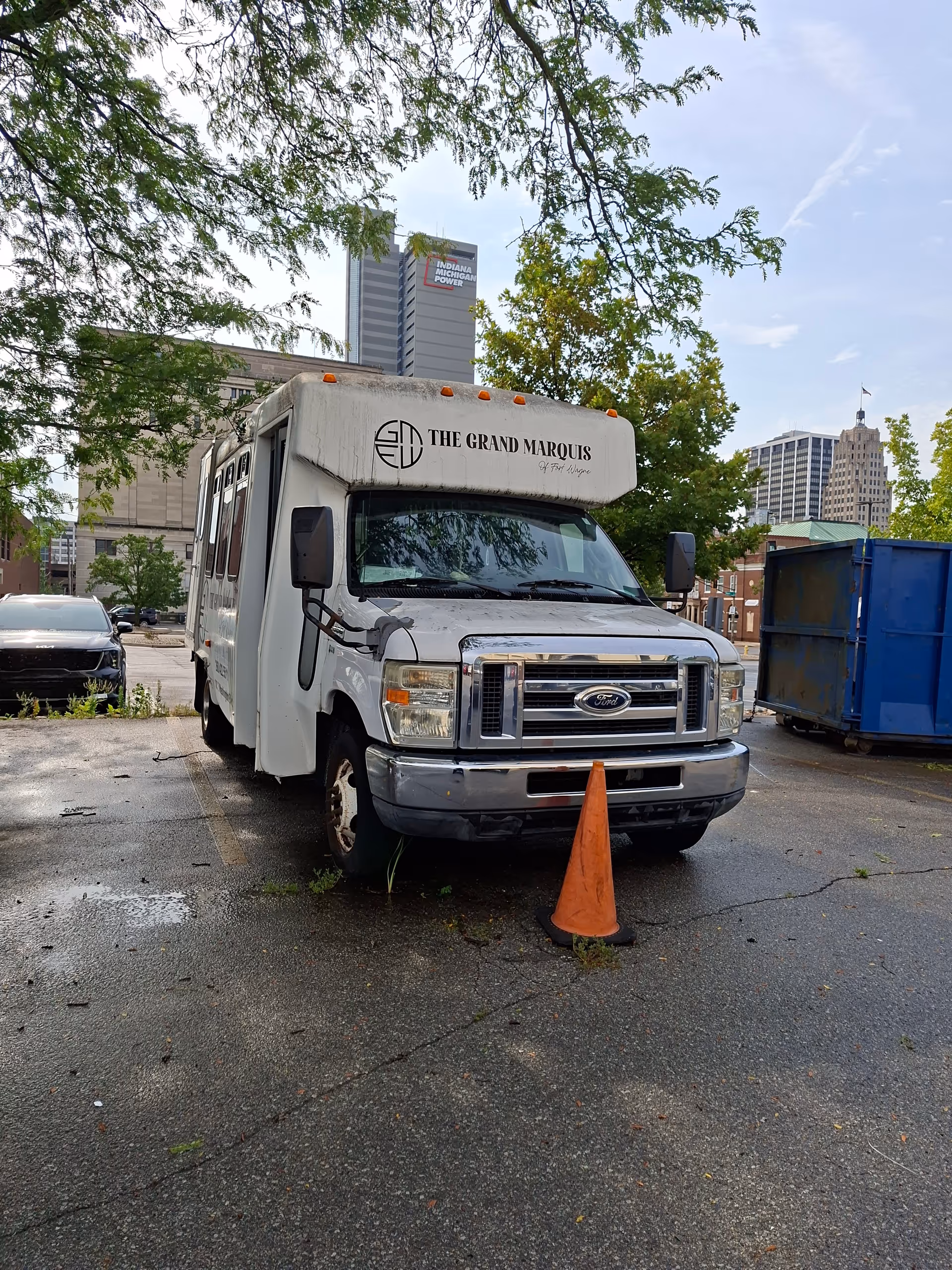 White shuttle bus labeled The Grand Marquis parked in a tree-lined, wet lot.