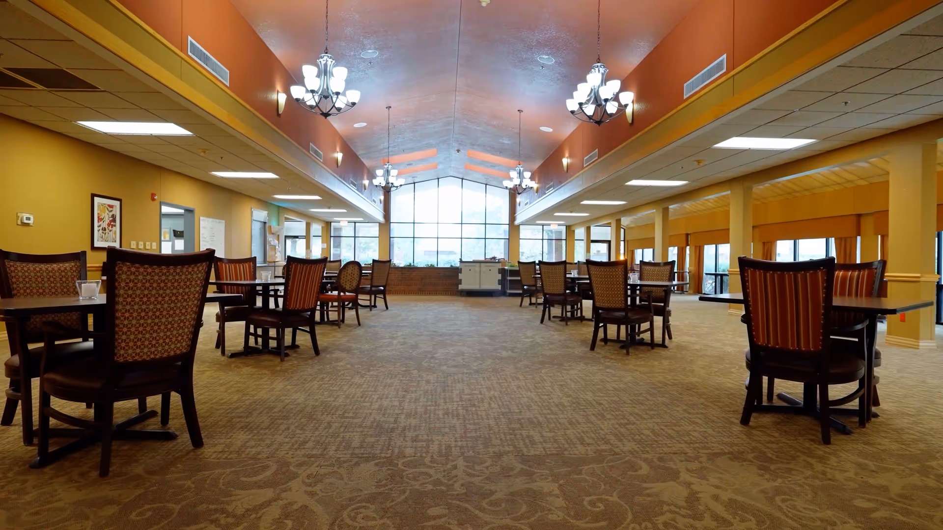 A spacious dining room with multiple tables and chairs arranged neatly. The room features warm yellow walls, large windows at the far end allowing natural light, and chandeliers hanging from the ceiling. The carpet has a subtle patterned design, and the overall atmosphere is inviting and comfortable.