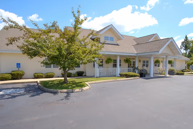 Exterior view of a single-story senior living facility building with a covered entrance, white siding, and a small tree in front. The sky is clear with a few clouds.