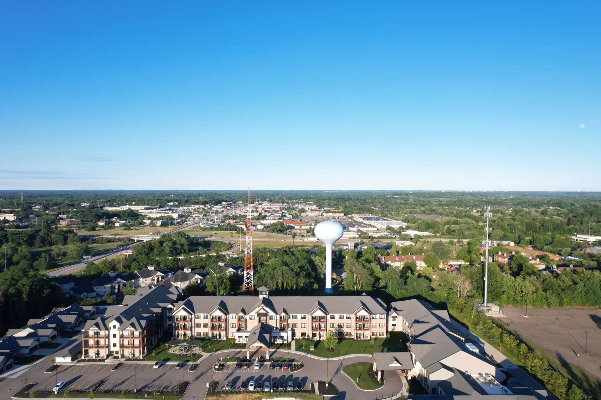 Aerial view of a large multi-story residential building complex with a parking lot in front, surrounded by greenery and other buildings in the distance under a clear blue sky.