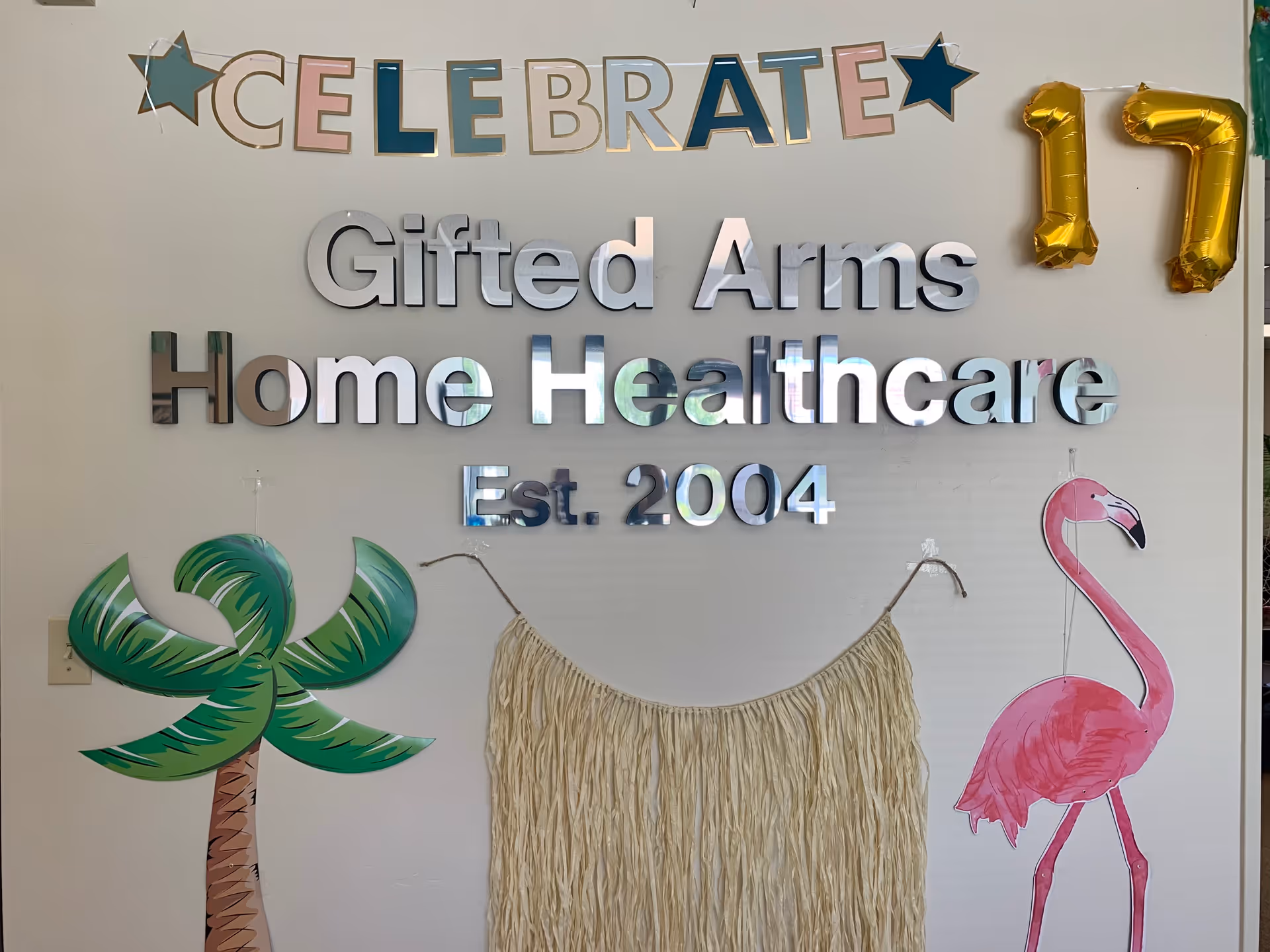 Indoor wall display reads Gifted Arms Home Healthcare with celebration decorations and a flamingo artwork.