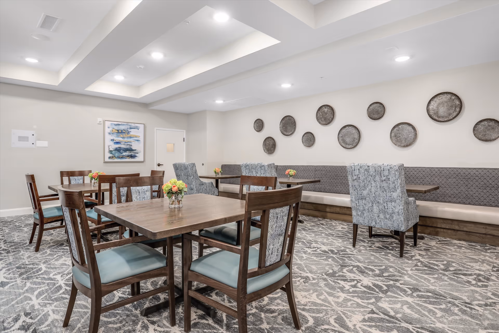 A dining room with wooden tables and chairs featuring blue cushioned seats. The room has a patterned carpet, a long cushioned bench along one wall, and decorative round wall hangings. Small floral arrangements are placed on each table. The ceiling has recessed lighting and a tray design.