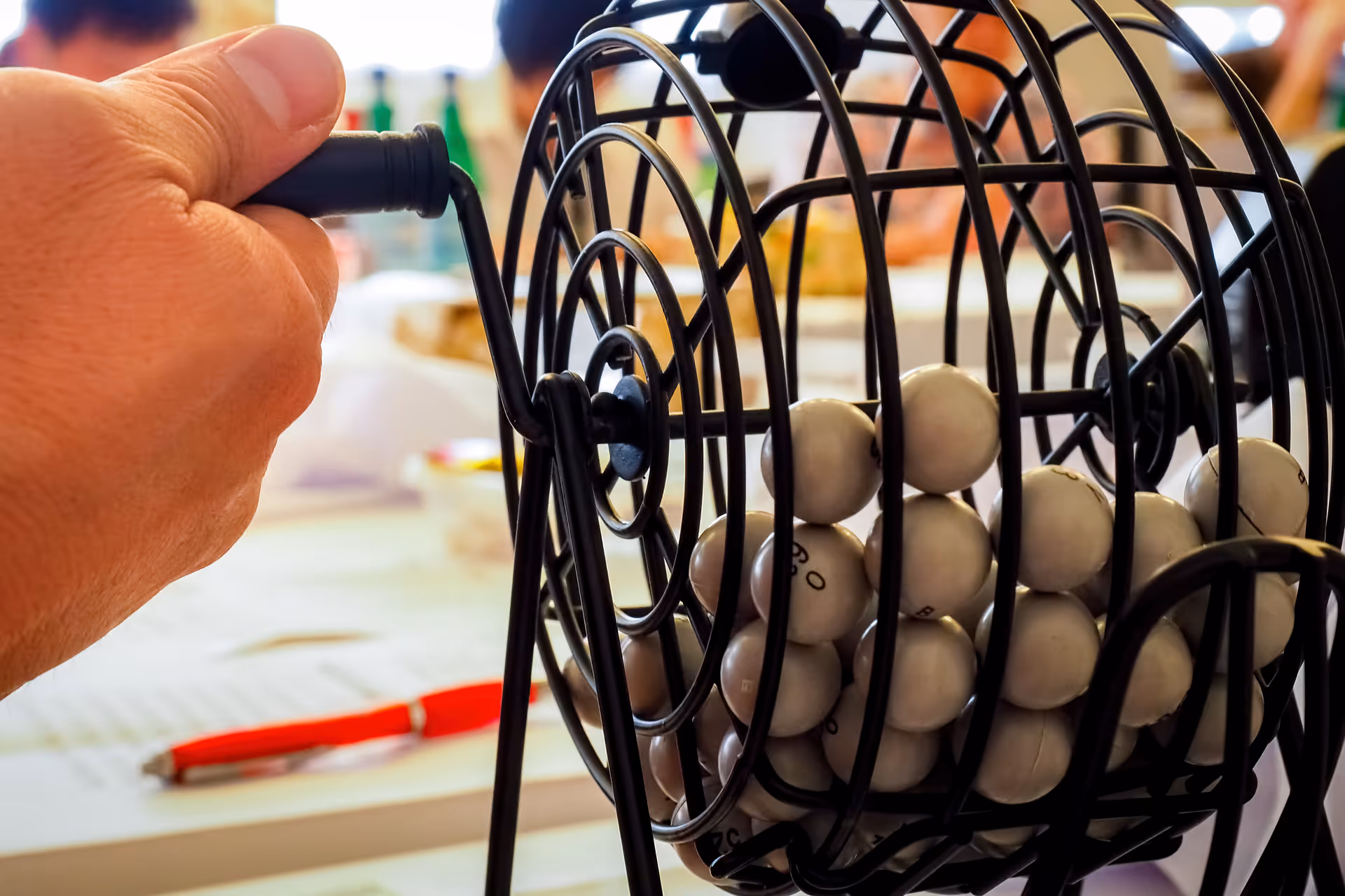 A hand turns a bingo cage filled with white balls on a table.