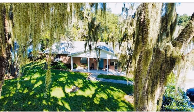 A single-story brick building with a covered porch featuring chairs, surrounded by green grass and large trees with hanging moss, under a clear sky.