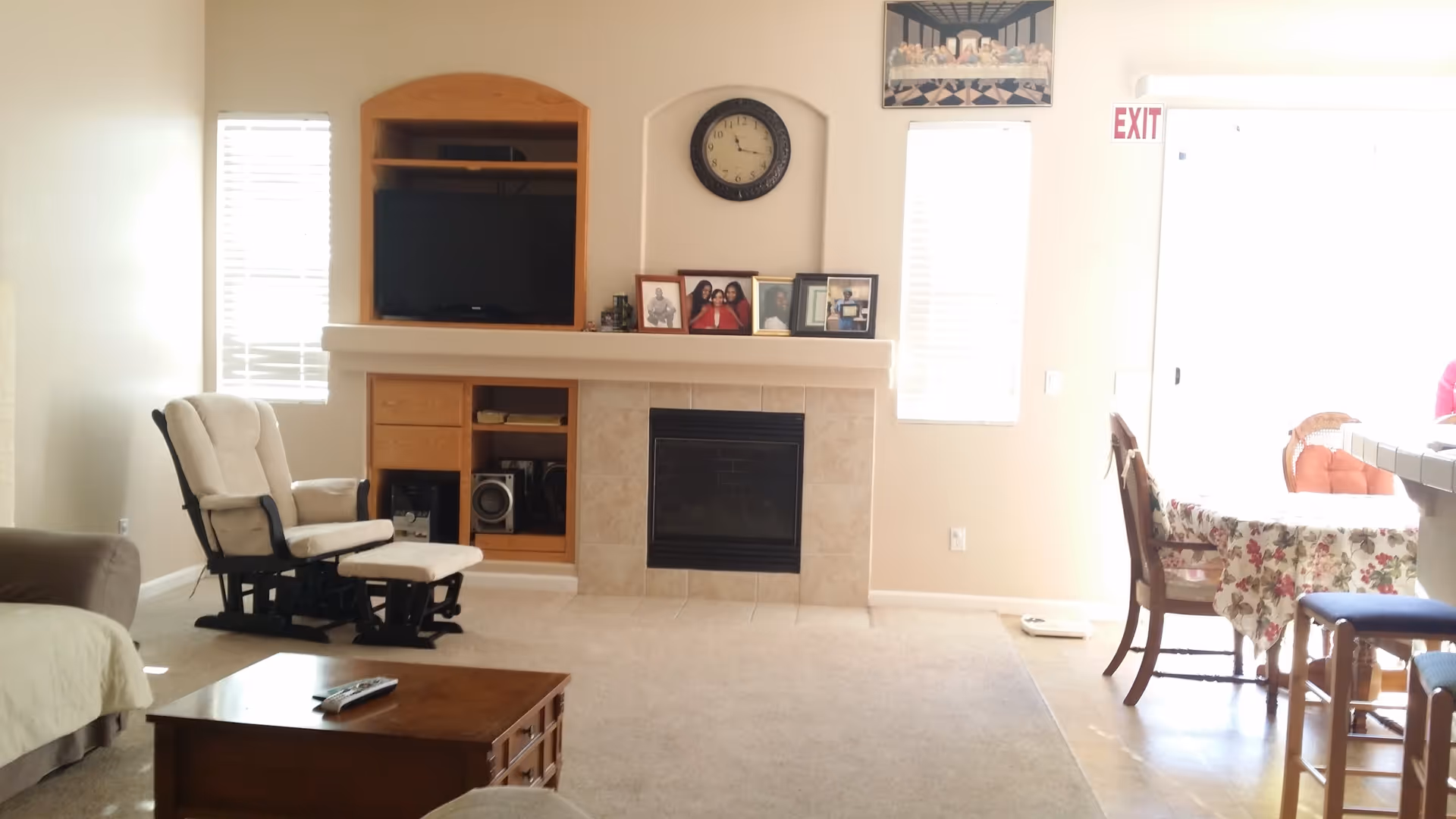 A bright living room with a beige carpet and light-colored walls. There is a fireplace with a tiled surround and a built-in wooden entertainment center above it, holding a TV and some electronic devices. On the mantel are framed photos and a round wall clock above. To the left is a cushioned rocking chair with an ottoman, and a brown sofa partially visible. To the right, there is a dining area with a floral tablecloth on the table and wooden chairs. Sunlight streams in through windows with blinds, and an exit door is visible in the background.