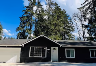Single-story dark building with white trim, front door, windows, and attached garage, trees behind.