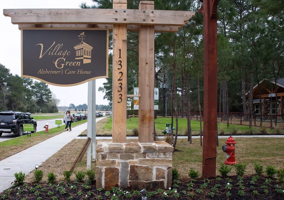 Wooden sign reading "Village Green Alzheimer's Care Home" mounted on posts at the facility entrance with a sidewalk, parked cars, trees, and a red fire hydrant visible.