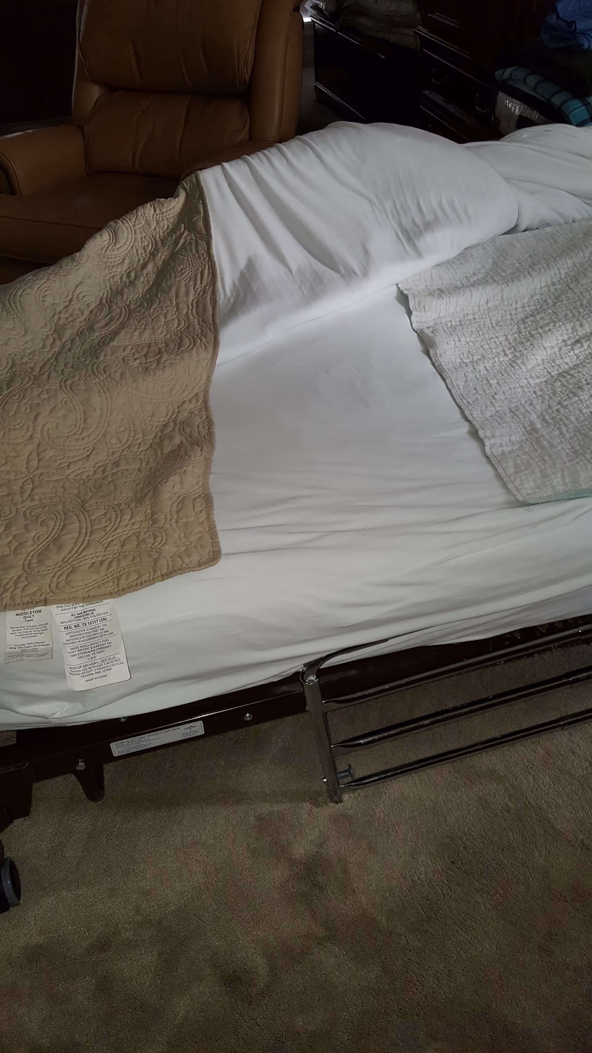 An adjustable bed with white sheets and beige quilt in a dim bedroom.