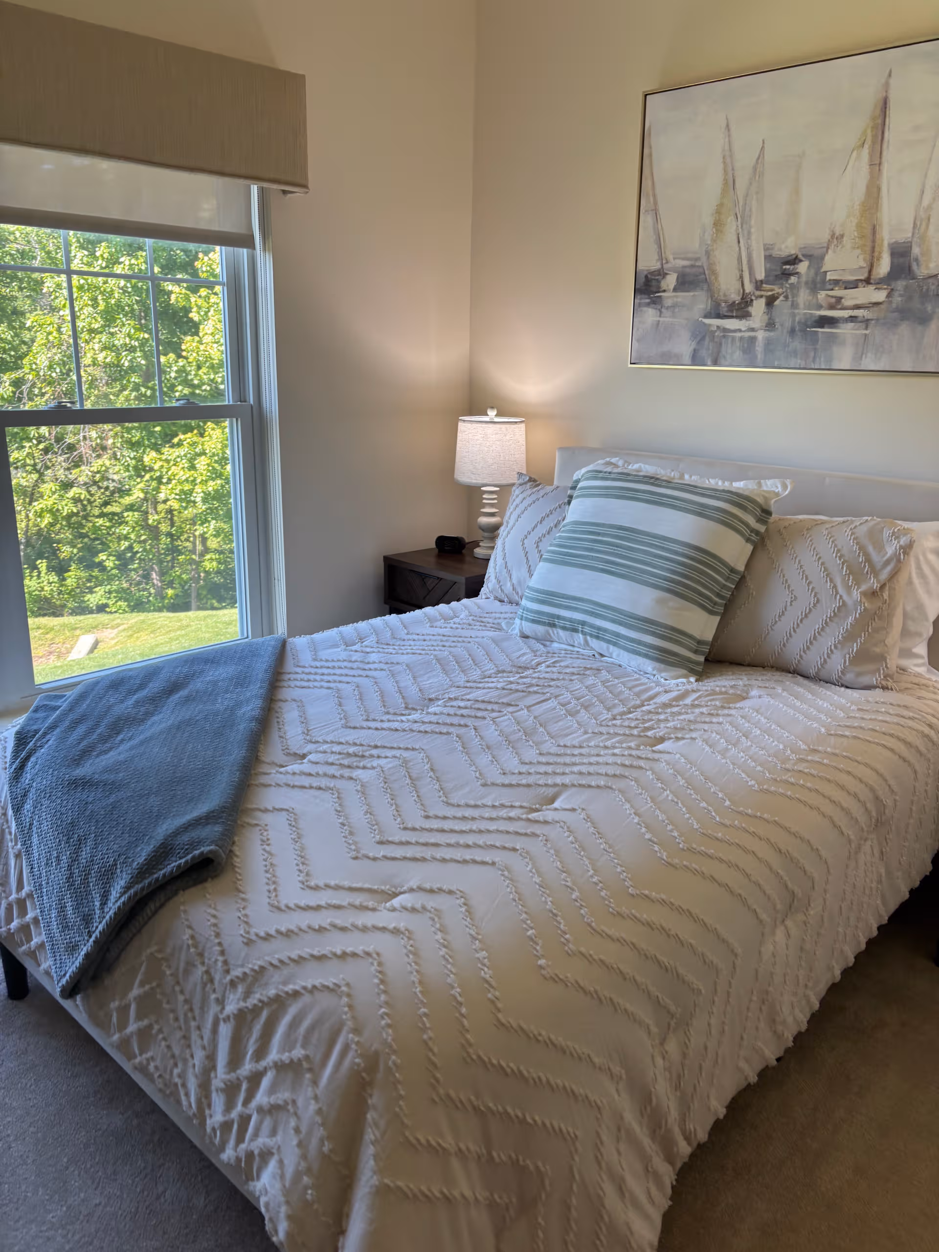 Bright bedroom with a white chevron quilt, striped pillows, nightstand, lamp, and a large window.
