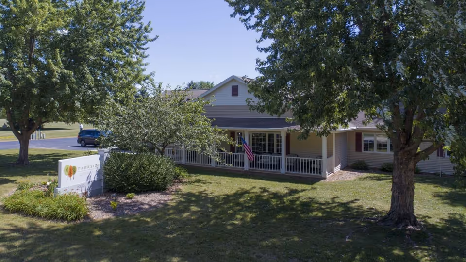Single-story senior living facility with a front porch, flag, and surrounding trees.